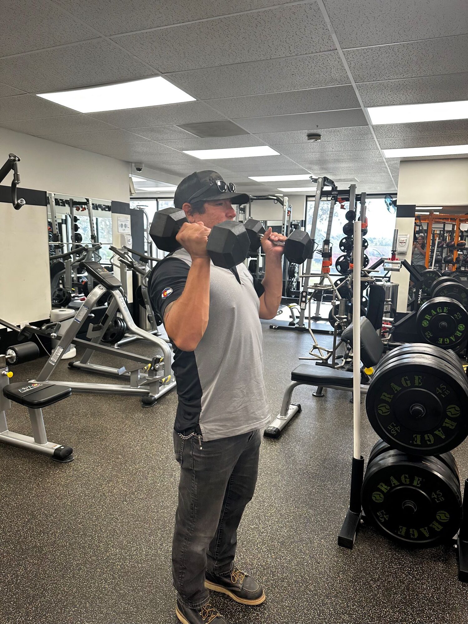 Man in black pants, gray shirt, and black baseball cap holds a dumbbell in each hand on his shoulders at AFAC gym