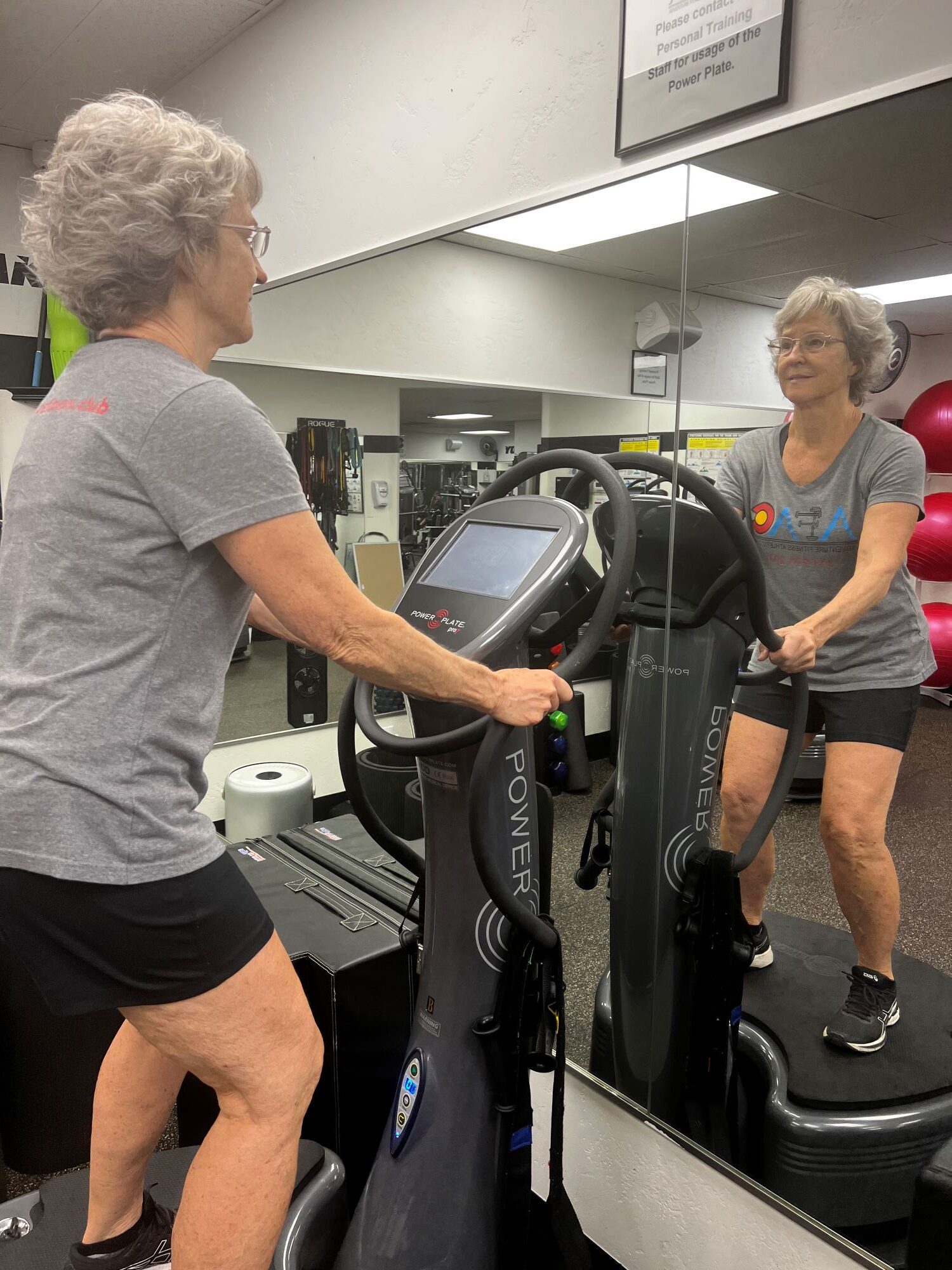Older woman in gray t-shirt and black shorts stands on a Power Plate at AFAC gym