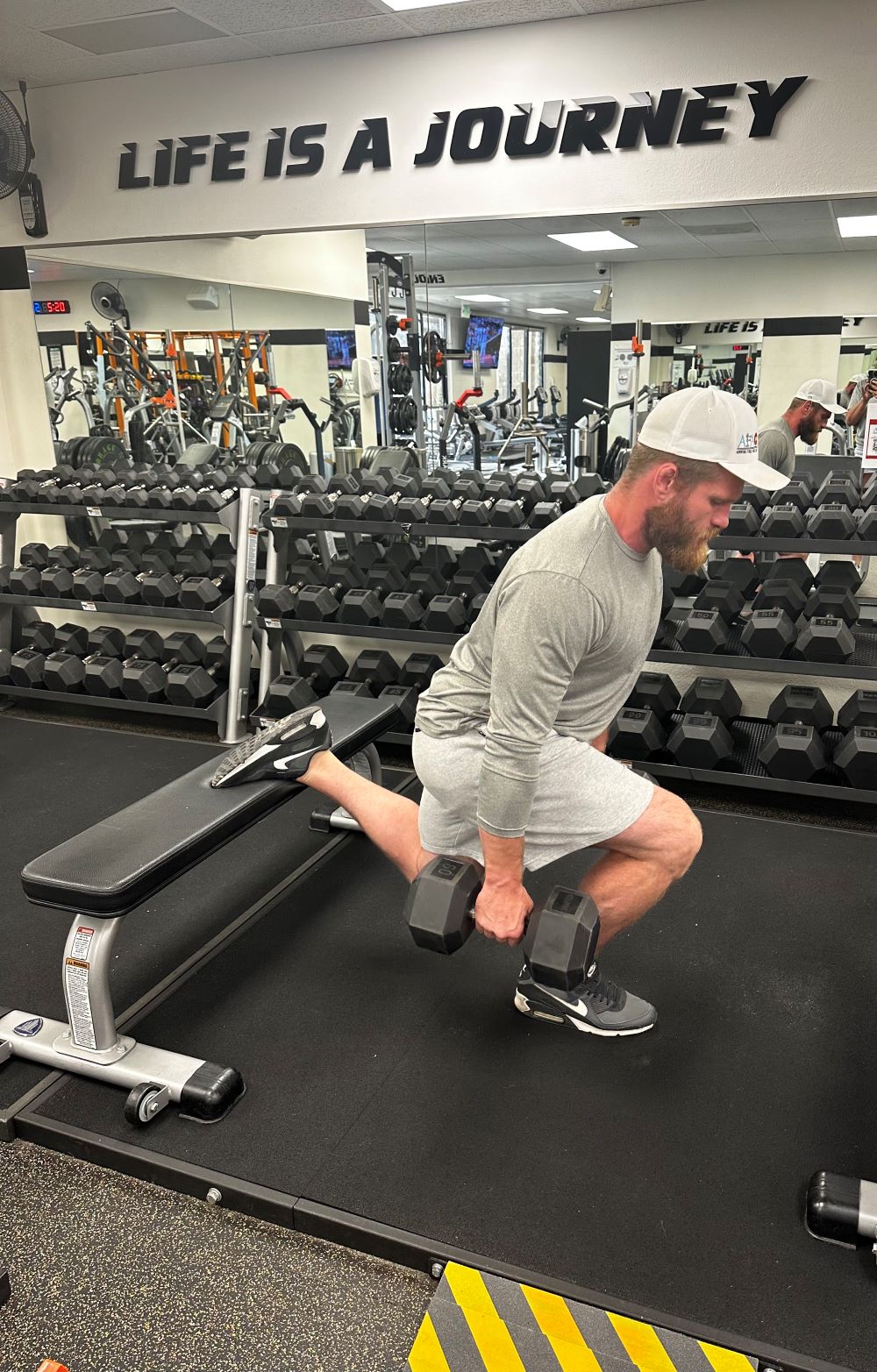 Man in white hat, gray shirt, and gray shorts performs Bulgarian split squat while holding dumbbells at AFAC gym