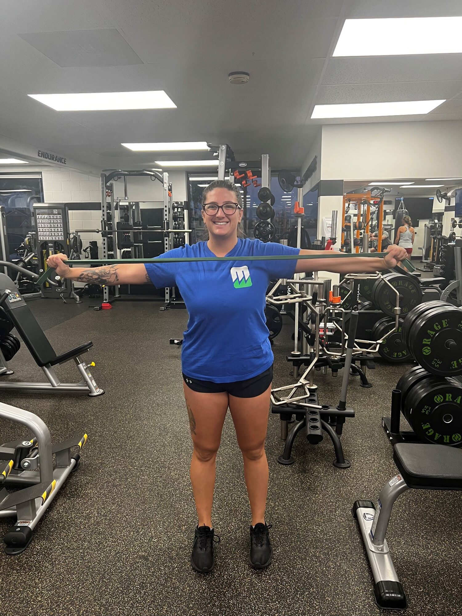 Standing woman in blue t-shirt and black shorts smiling and working out with a resistance band at AFAC gym