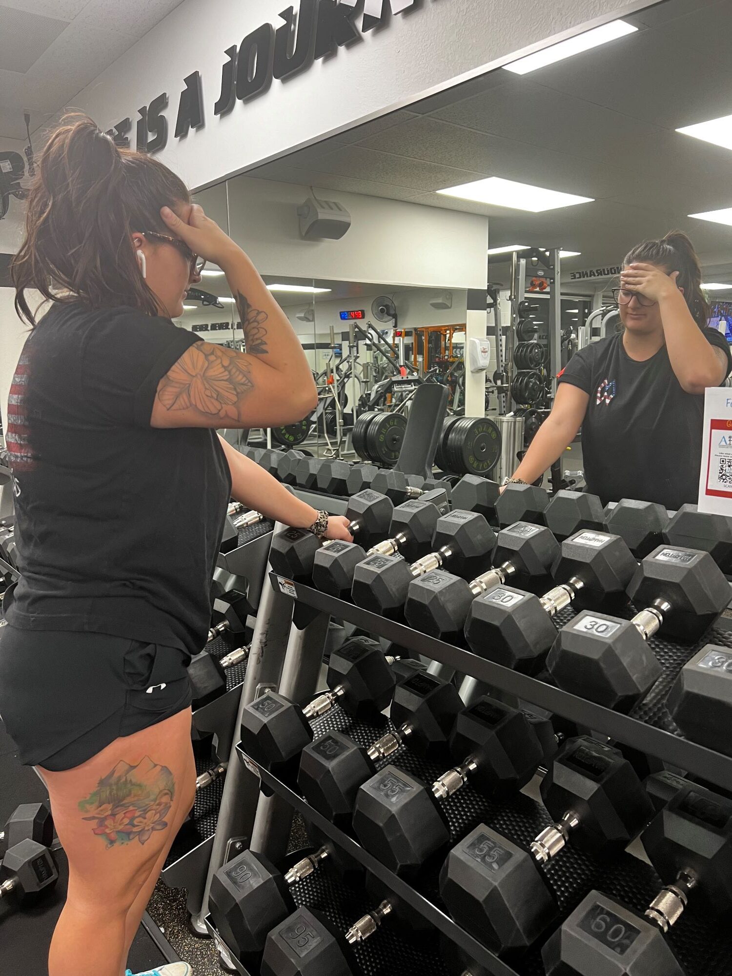 Woman dressed in a black shirt and shorts at AFAC gym. She's standing in front of a dumbbell rack with her hand on her forehead.