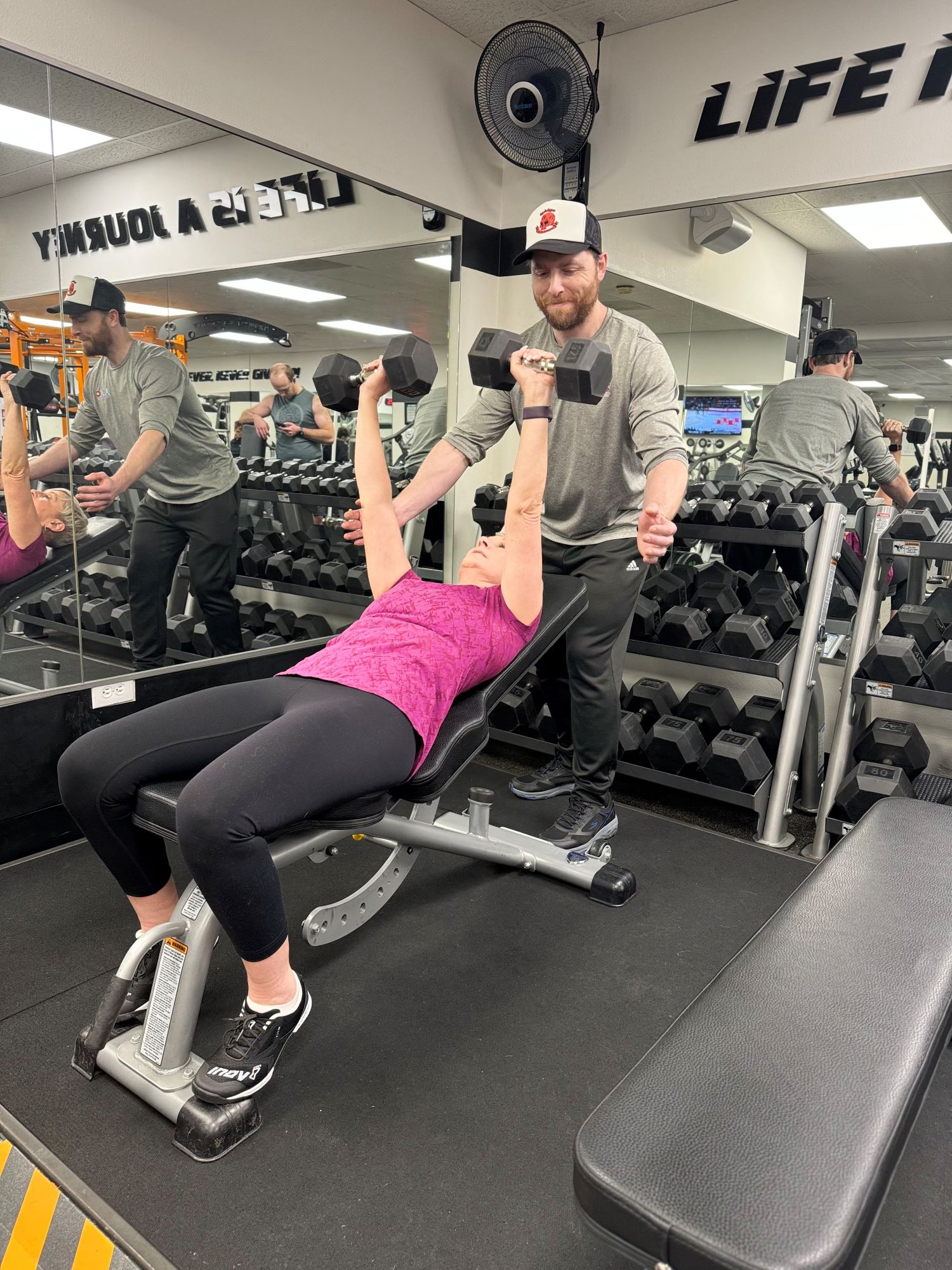 Woman laying on bench lifting two dumbbells with the assistance of a personal trainer at AFAC gym