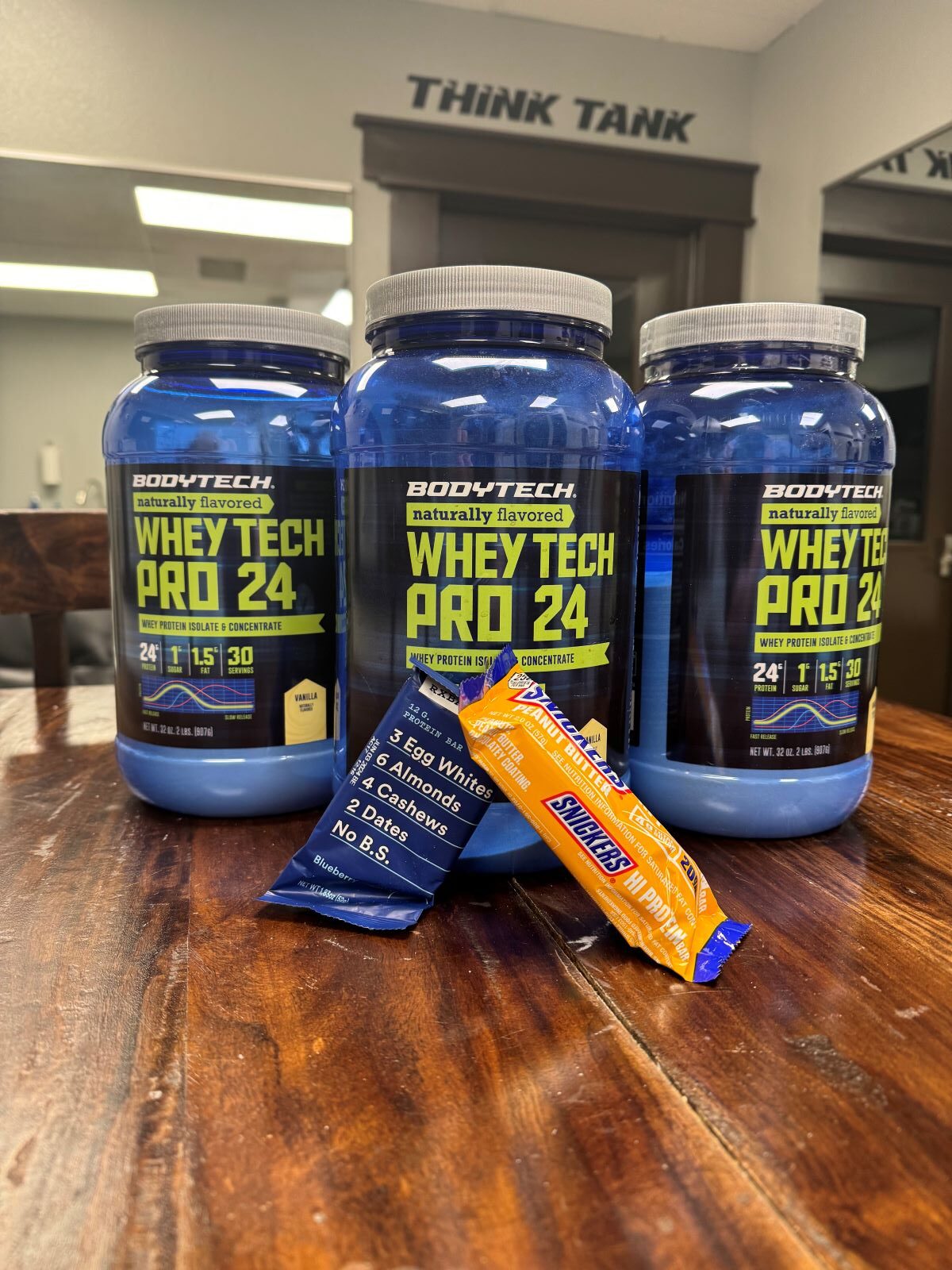 Three tubs of vanilla whey protein powder, a Snickers protein bar, and blueberry protein bar on a table at AFAC gym