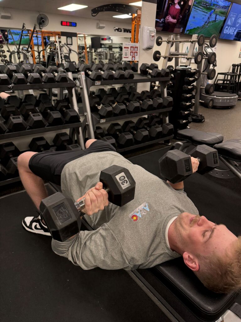 Man laying on a bench at the gym holding two dumbbells by his chest