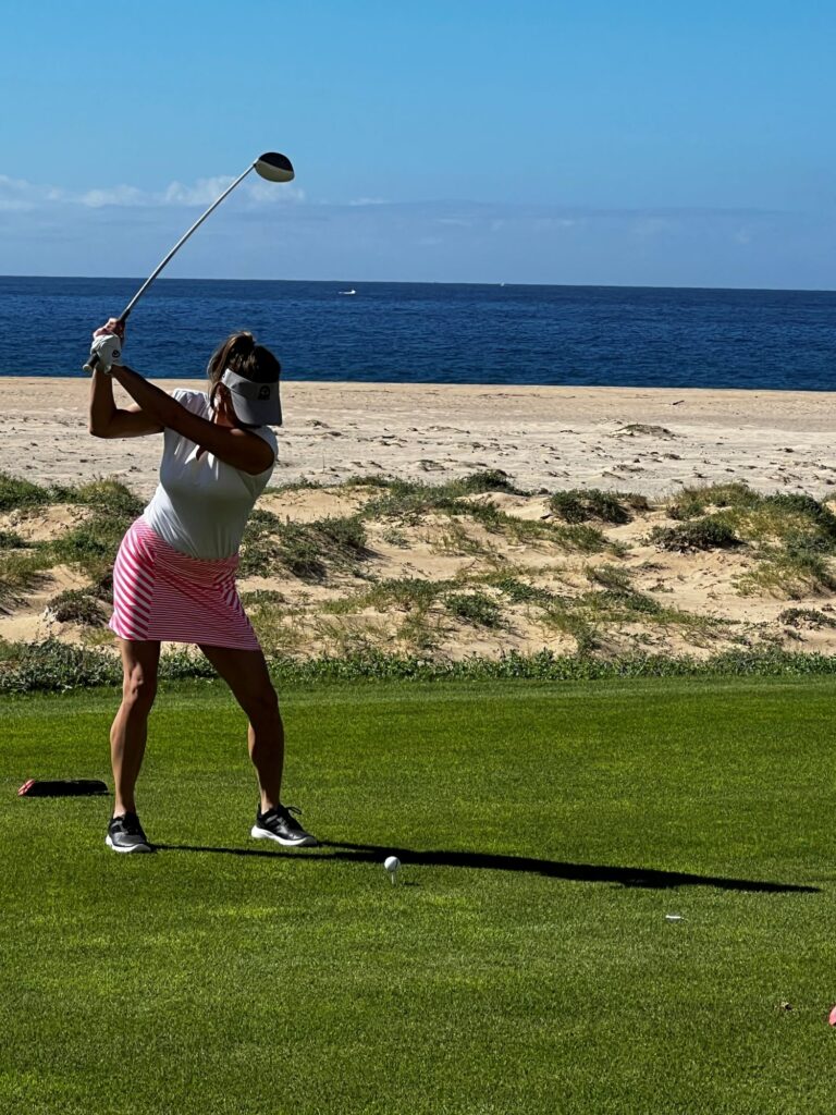 Woman swinging golf club on golf course with beach and water behind her