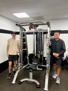 Two smiling men standing by a squat rack at AFAC gym