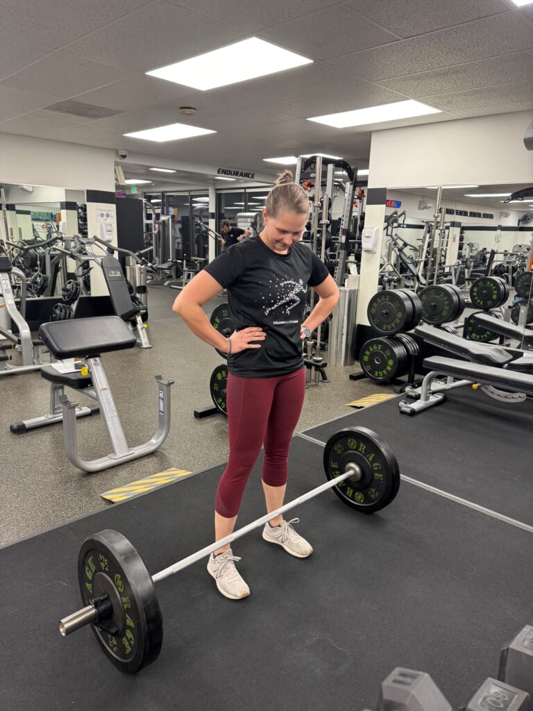 Woman standing with her hands on her hips, looking down at a loaded barbell on the floor