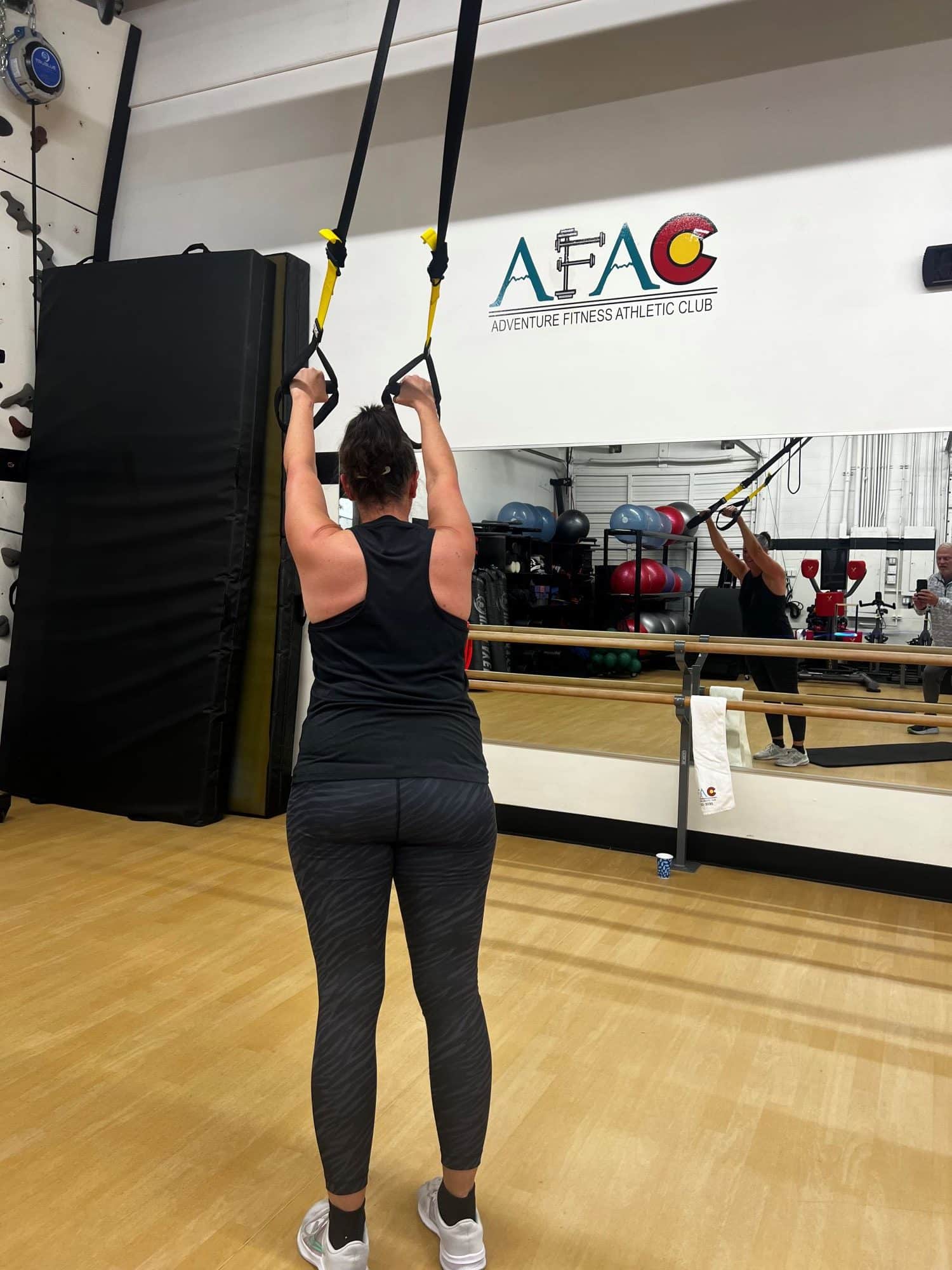 Back view of woman at AFAC gym standing up and exercising with a TRX suspension trainer with her arms overhead