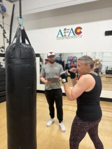 Woman works out during her class schedule with punching bag at AFAC gym while a male personal trainer looks on holding a clipboard
