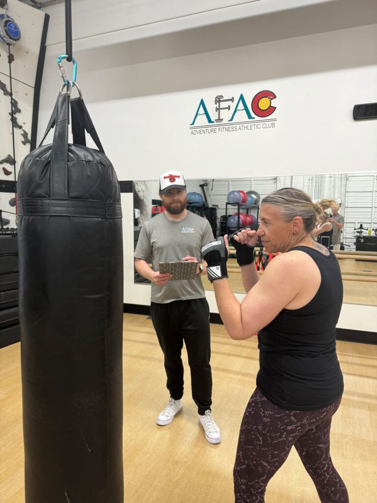 Woman works out during her class schedule with punching bag at AFAC gym while a male personal trainer looks on holding a clipboard