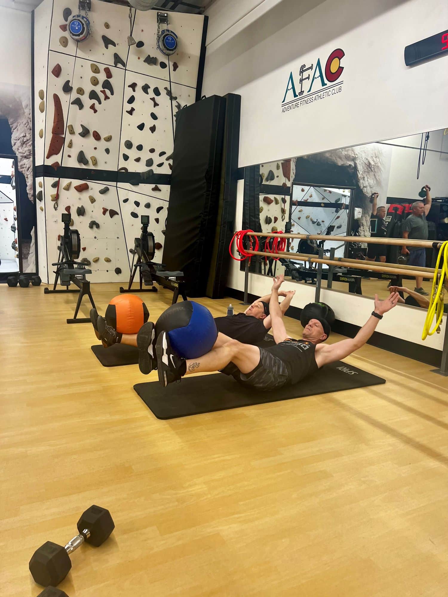 Two men lay down on exercise mats at AFAC gym while balancing exercise balls on their feet