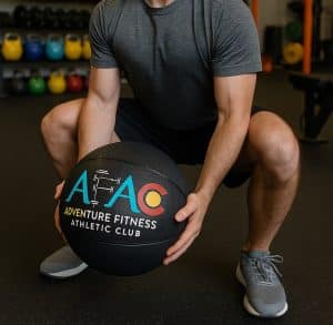 Man in shorts and a t-shirt squatting down and holding an AFAC slam ball in front of him at AFAC gym