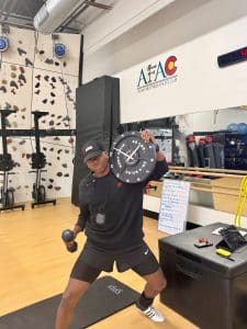 Personal trainer at AFAC gym holds a clock in one hand and a dumbbell in the other