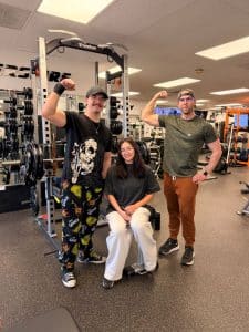 At AFAC gym, two men stand with right arms flexed while woman sits between them on a gym machine