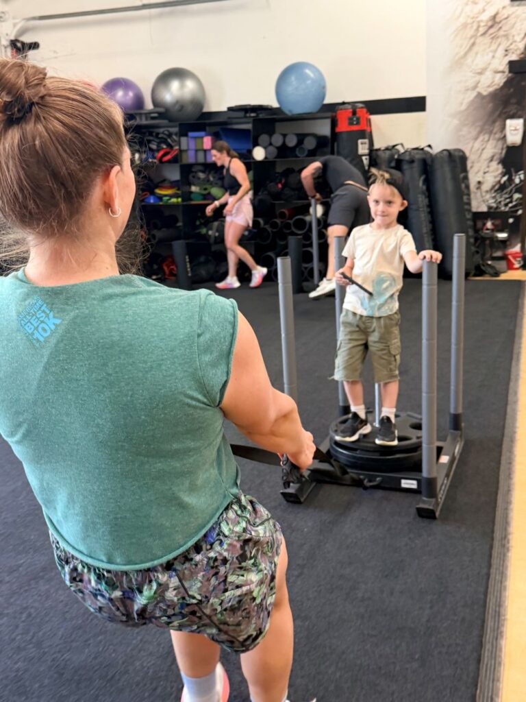 Woman pulling small boy on a gym sled over the gym turf area at AFAC gym