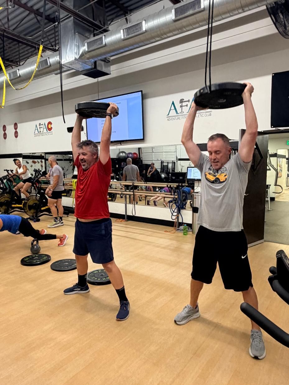 Two standing men holding weight plates above their heads during a circuit training class at AFAC gym
